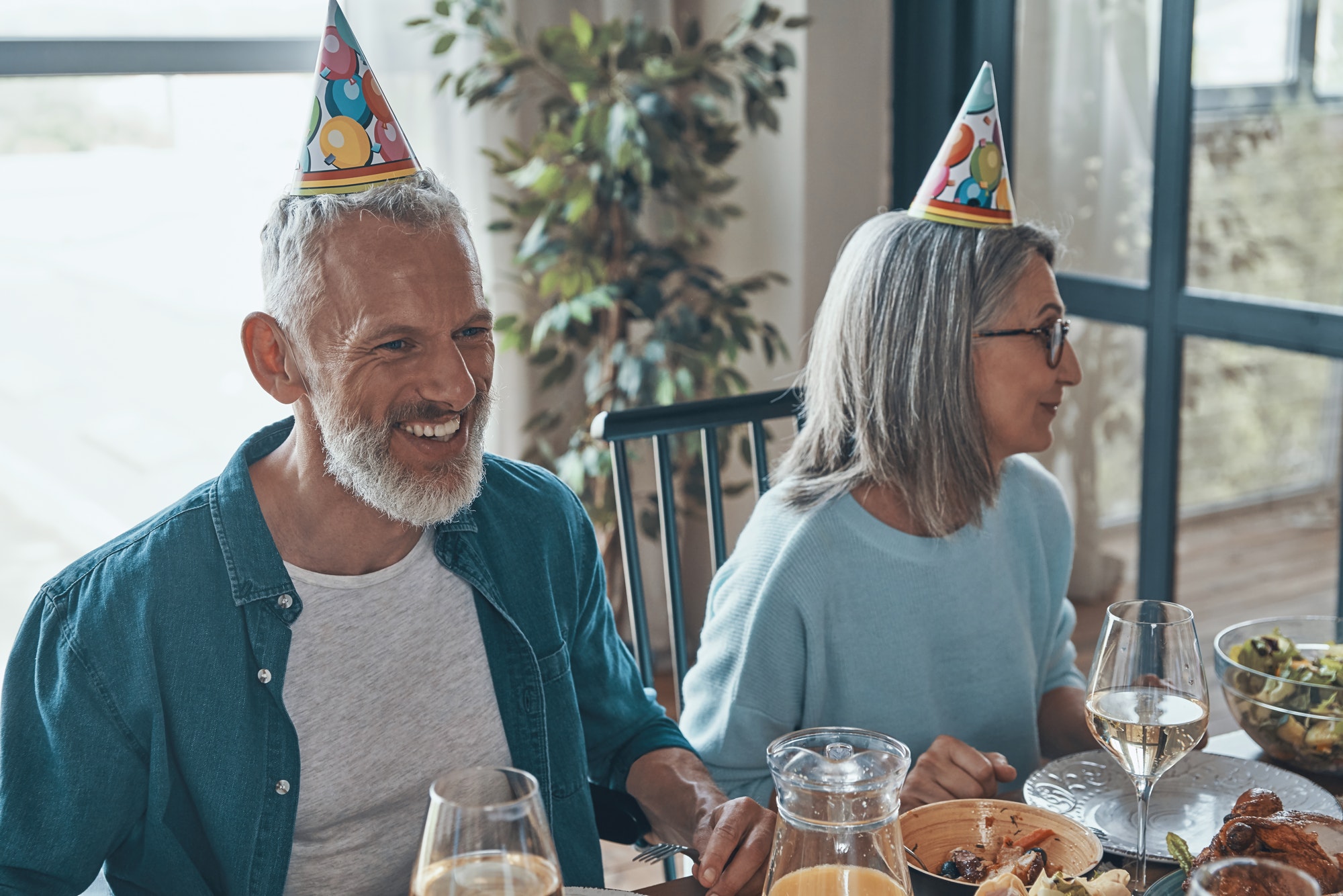 Happy senior couple in funky hats celebrating birthday while sitting at the dining table at home after enrolling in Medicare Part D in the state of Vancouver